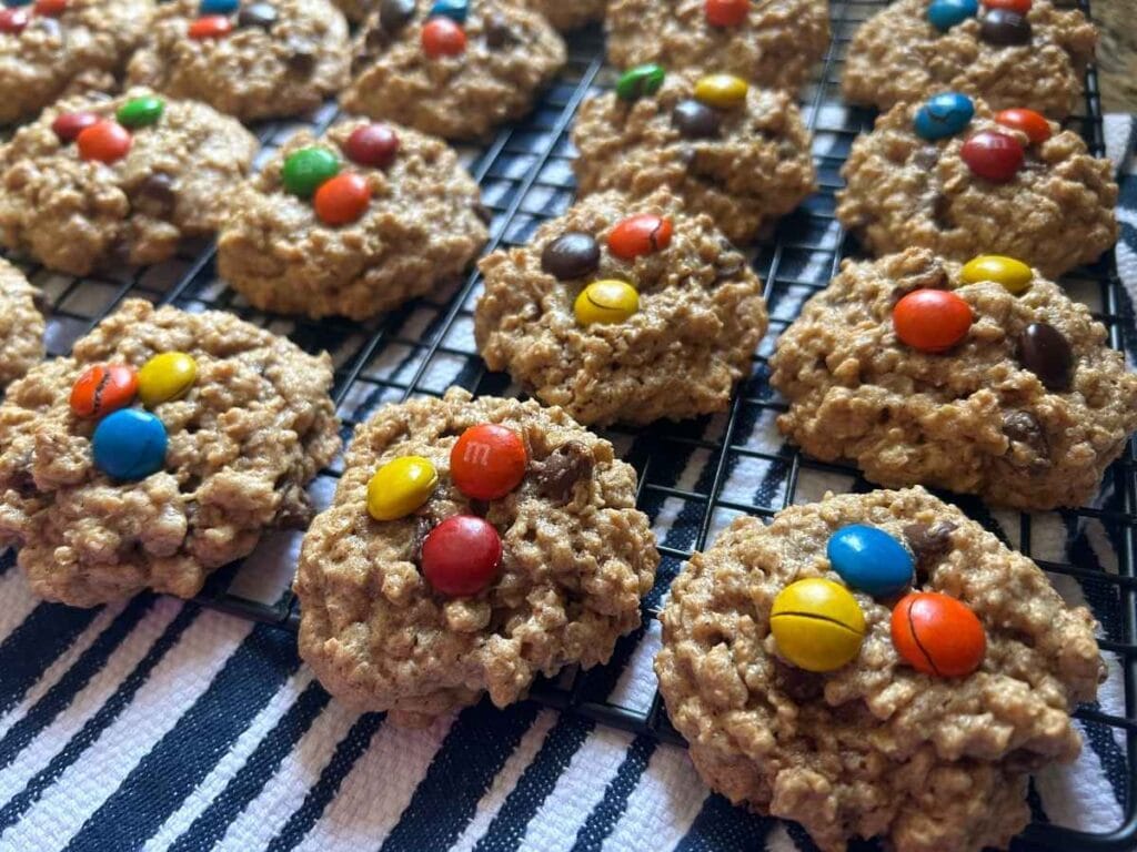 oatmeal chocolate chip cookies with m&ms on top cooling on a rack over a navy stiped towel
